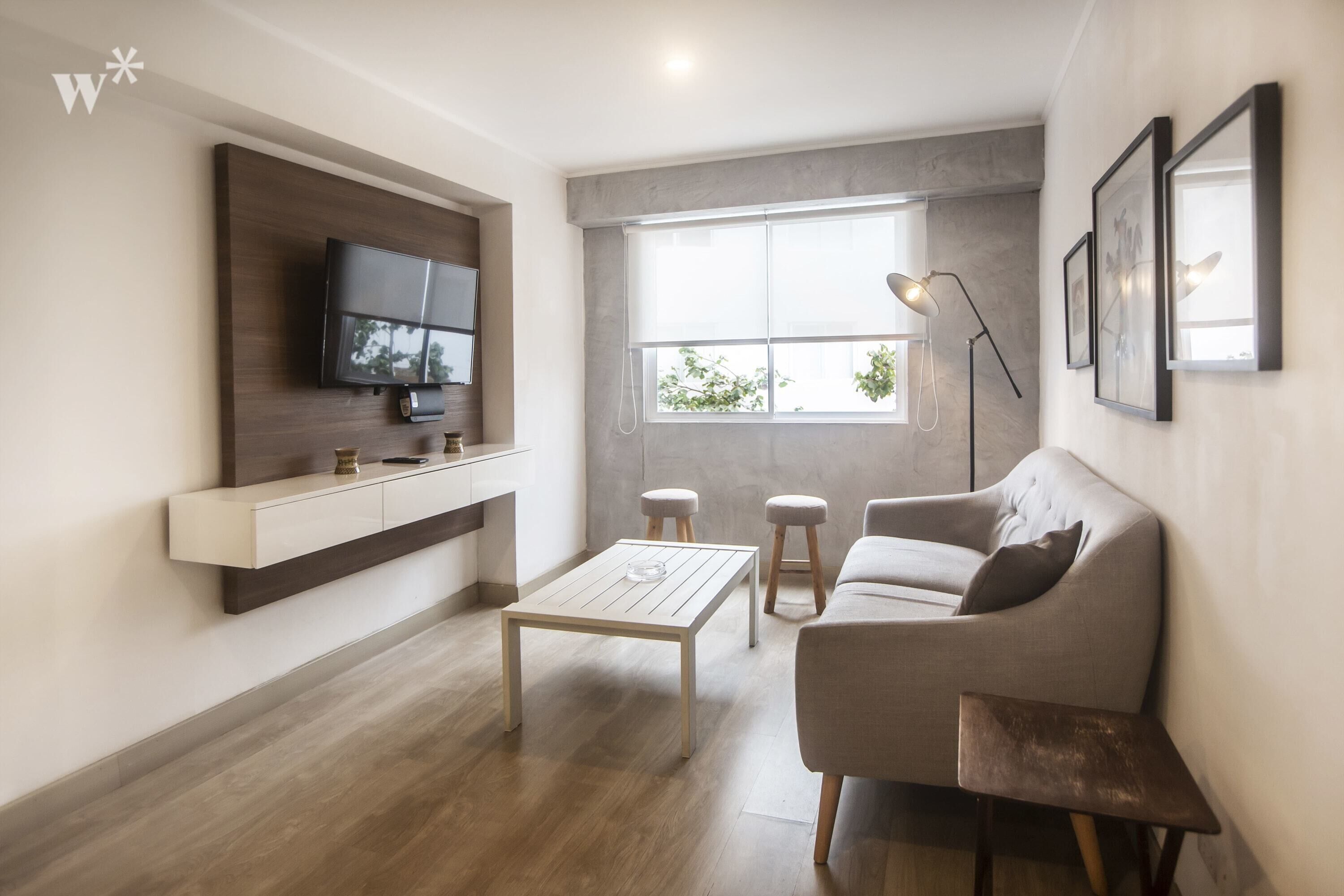 Ein modernes Wohnzimmer mit einem an einer Holzvertäfelung montierten Flachbildfernseher, einem Holztisch, einem grauen Sofa, zwei hölzernen Hocker und einer Stehlampe.