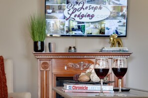Ett vardagsrum med en öppen spis, en tv-skärm som visar ”Joseph Lodging.com” och två glas rött vin på ett bord tillsammans med några böcker.