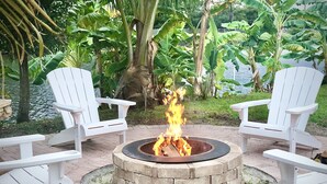 A circular fire pit with burning logs surrounded by white Adirondack chairs, set on a brick patio with a view of a water body and lush greenery. - Make yourself at home in Delray Beach!!<br><br><br> (Delray Beach)