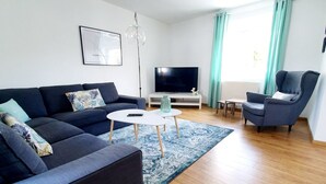 Una sala de estar moderna con un sofá oscuro, una pequeña mesa de centro, un televisor sobre un mueble blanco y una silla azul.