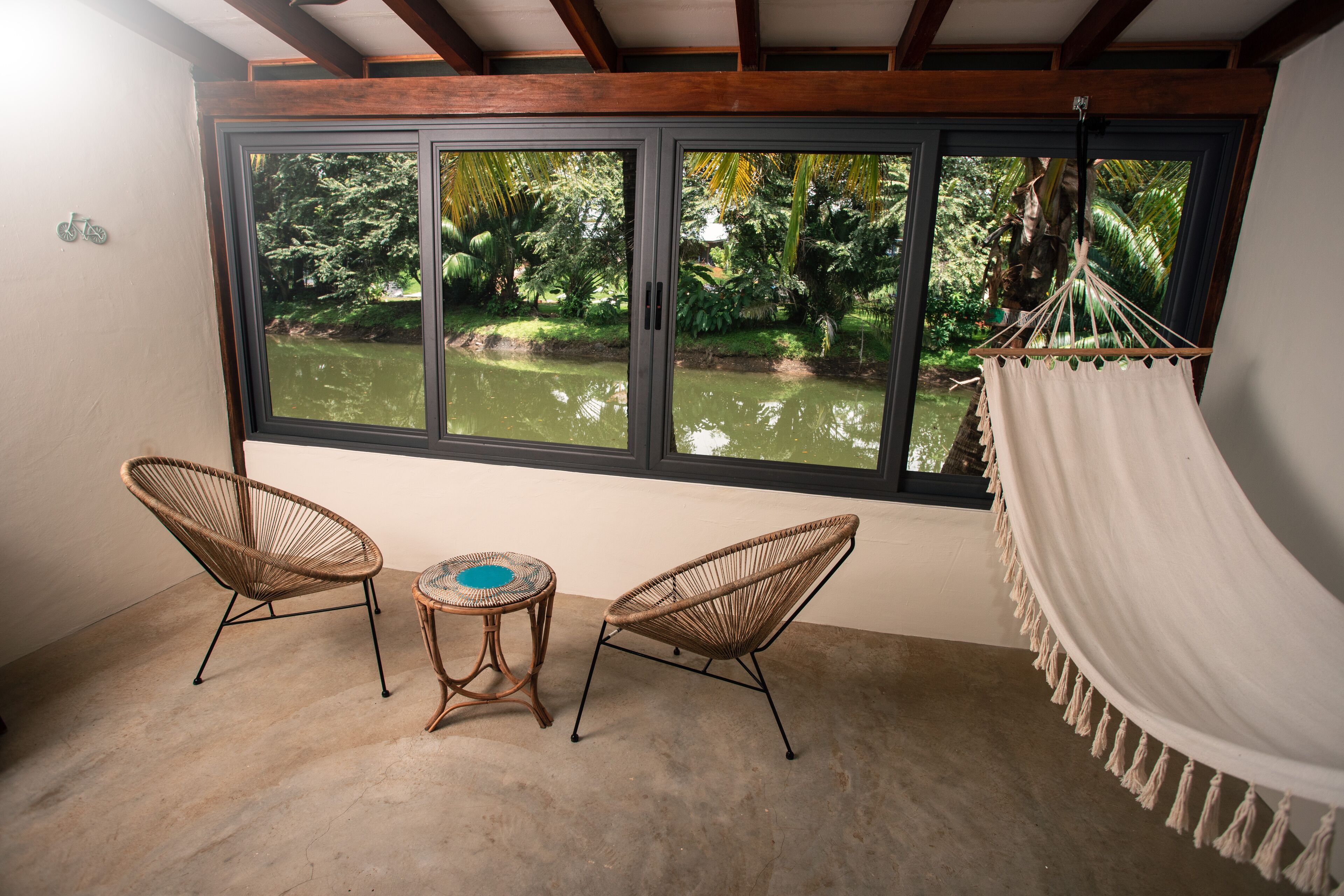 A room with a large window, two wicker chairs, a small round table, and a white hammock.