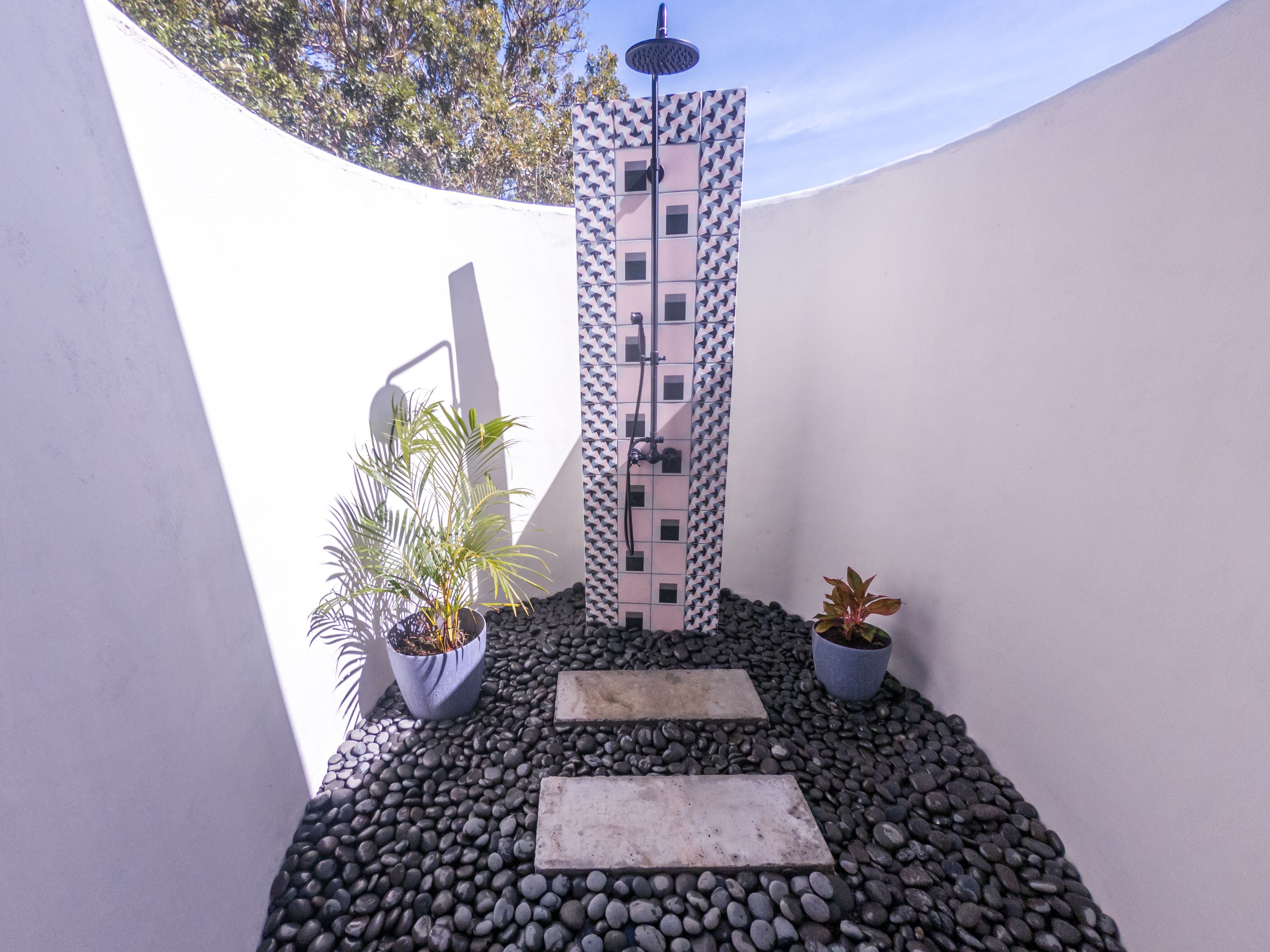 A modern outdoor shower area with a patterned wall, potted plants, and a pebble ground.