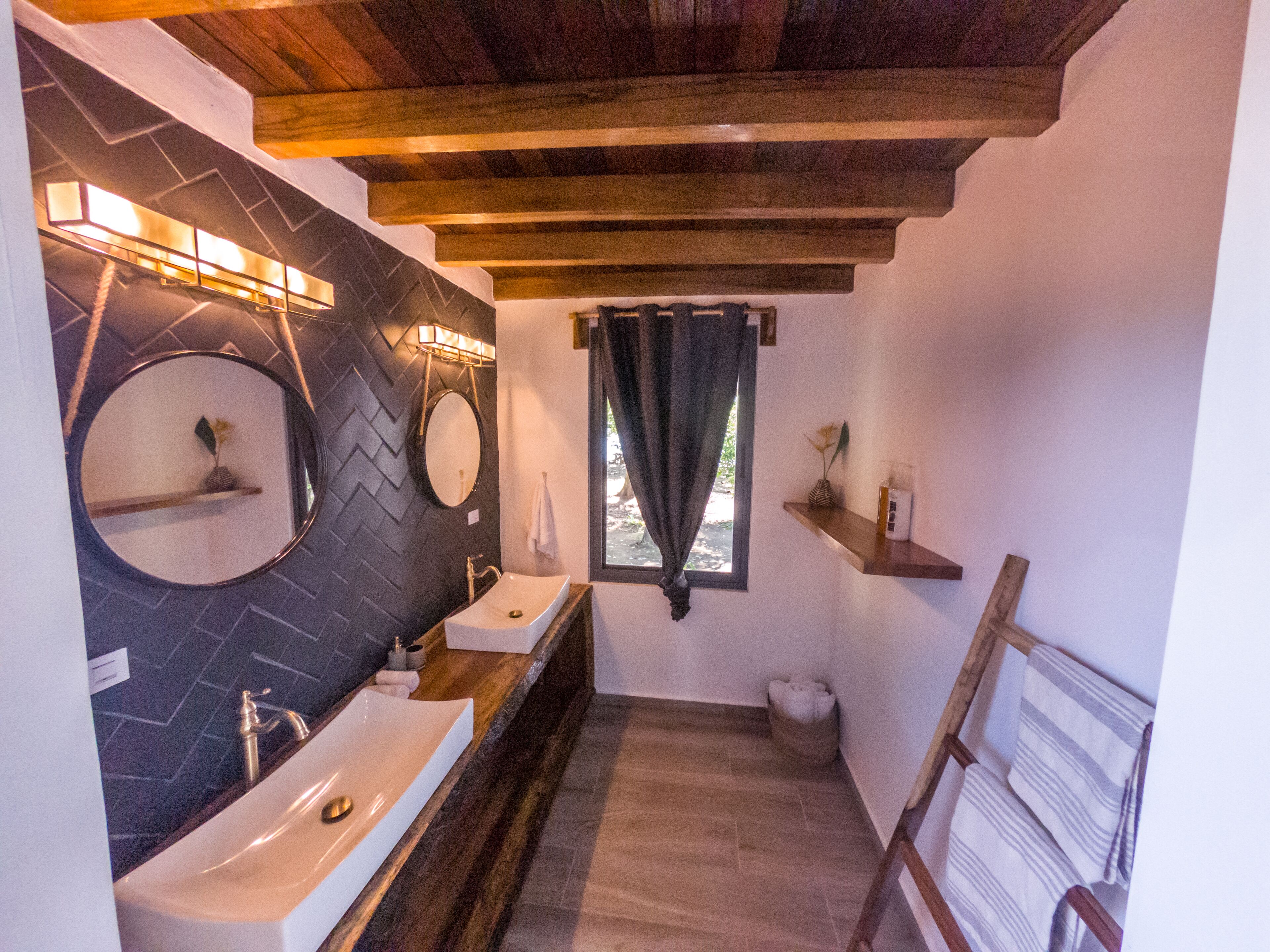 Un baño con techo de madera, un espejo grande, dos lavamanos y un ventanal con cortinas.
