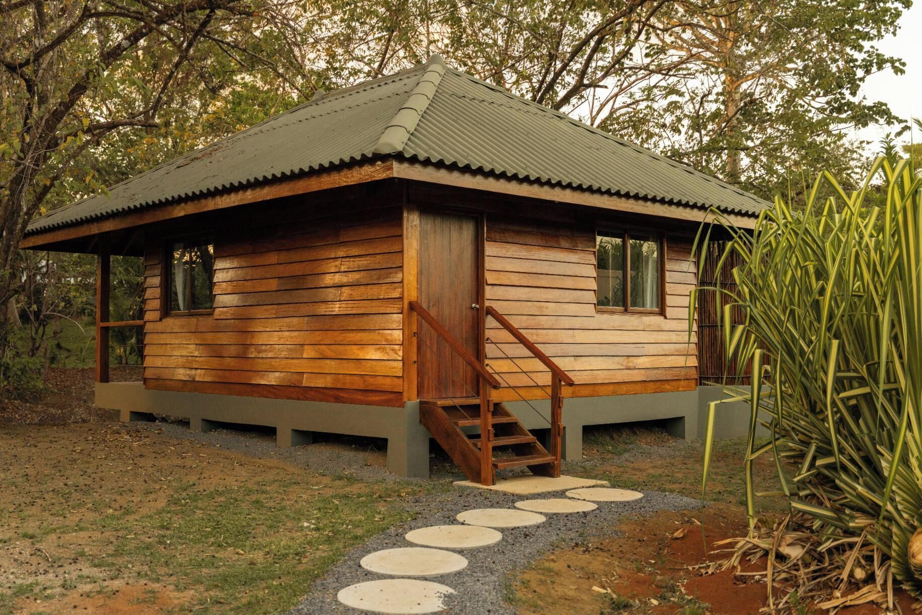 Une cabane en bois avec un toit en tôle ondulée, un escalier en bois et un chemin circulaire en pierre menant à l’entrée.