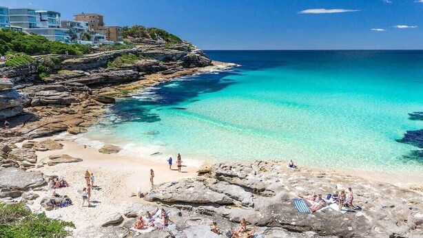 A coastal beach with rocky terrain, clear turquoise waters, and buildings in the background.