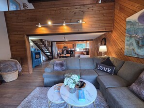 A cozy living room with a grey sectional sofa, a round coffee table with a vase of flowers, and a view into the kitchen and dining area.