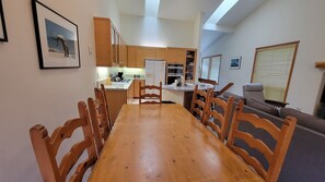 Een eetgedeelte met een houten tafel en stoelen, een keuken op de achtergrond, en een raam met zonwering.