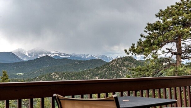 A balcony with a table and chairs, overlooking a mountainous landscape with snow-capped peaks and dense forest. - Work Away From Home- Mountain Home!!! Mountain Views of RMNP,Hot Tub,Sauna, Rural, Mountain Getaway-Top Of The World, Estes Park CO (Estes Park)
