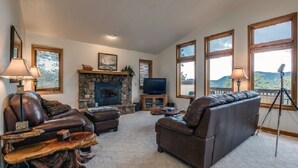 A spacious living room with a stone fireplace, leather sofas, and large windows offering a view of the outdoors. - Work Away From Home- Mountain Home!!! Mountain Views of RMNP,Hot Tub,Sauna, Rural, Mountain Getaway-Top Of The World, Estes Park CO (Estes Park)