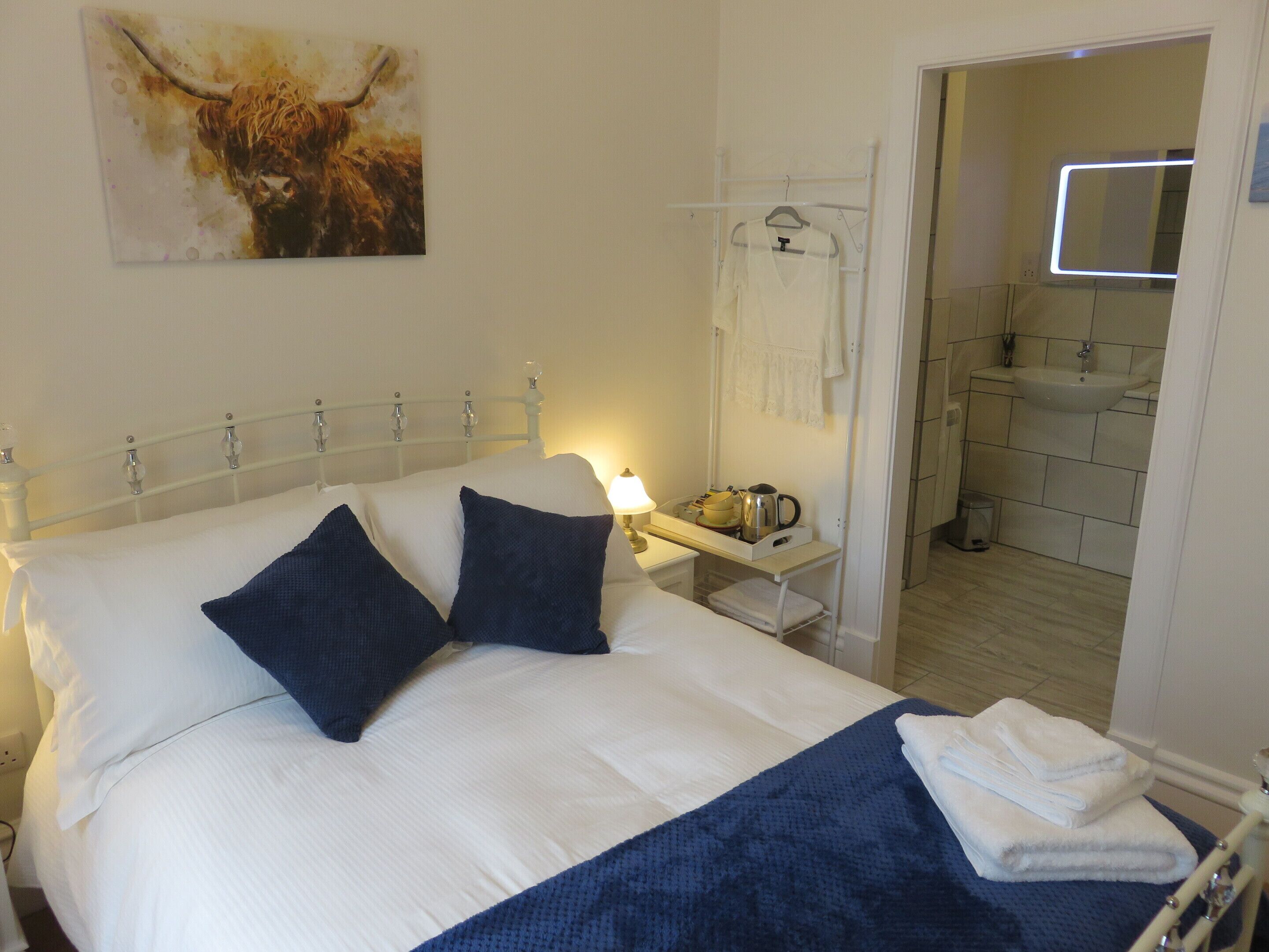 A neatly made bed with white and dark blue bedding, a bedside table with a lamp and a tray of items, and a painting of a bull on the wall.