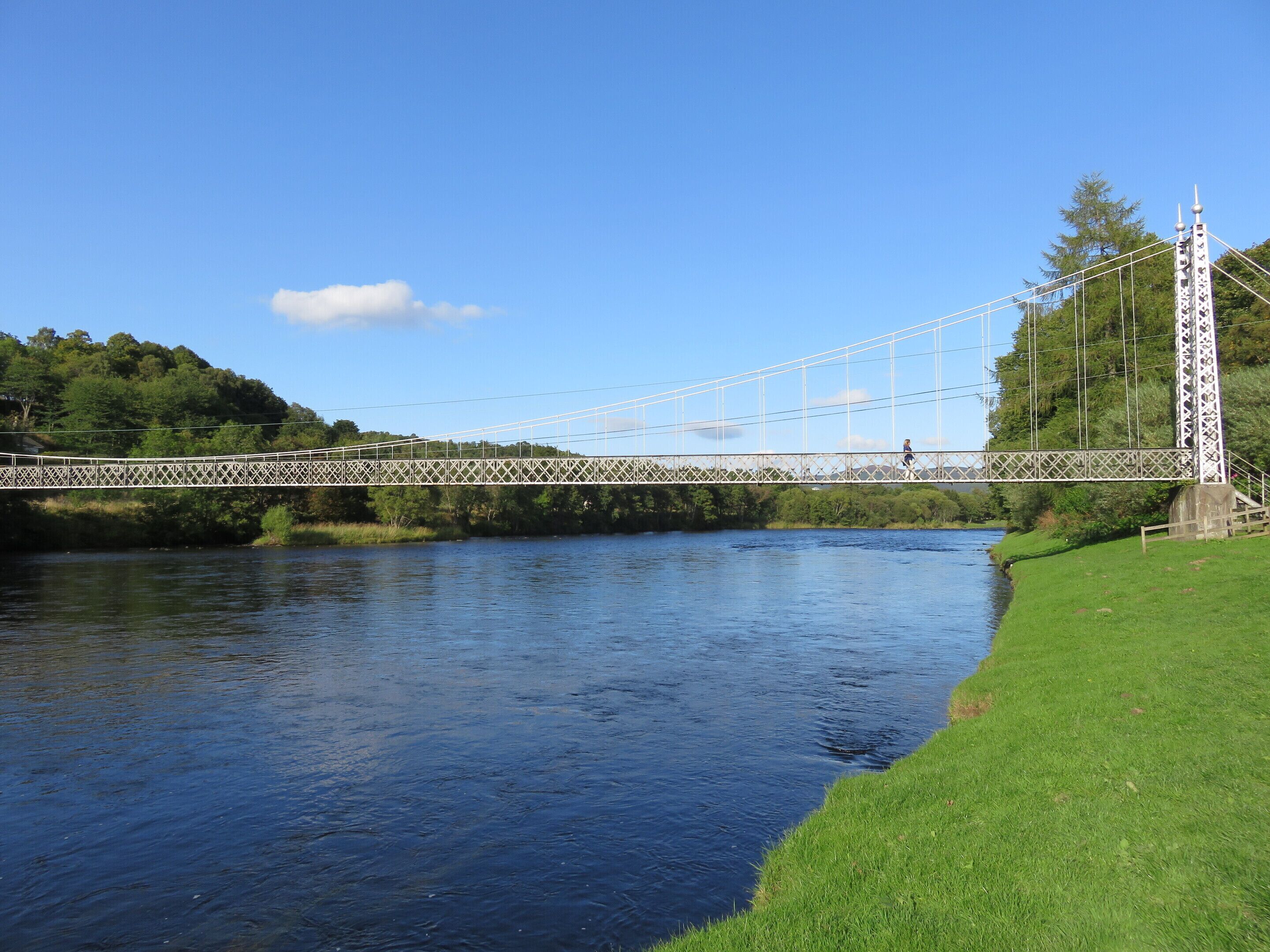 A suspension bridge over a river with a grassy bank and trees in the background.