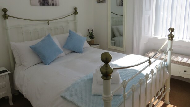 A neatly made bed with white bedding and blue pillows, a bedside table with a lamp, and a mirror on the wall. - Bankhouse B&B (Aberlour)