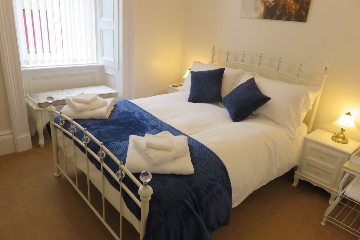 A neatly made bed with white and blue bedding, a bedside table with a lamp, and a window with blinds.