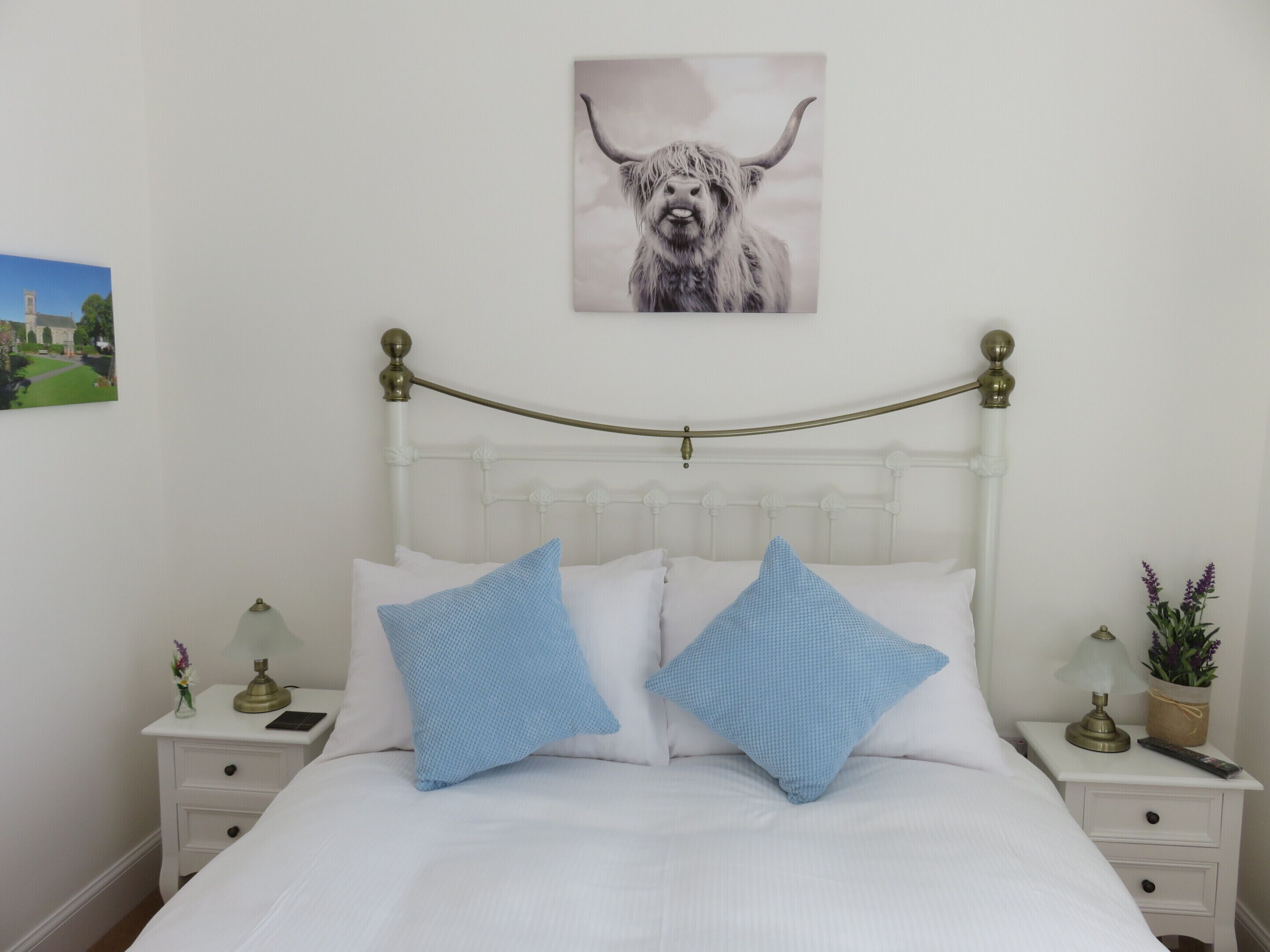 A neatly made bed with white bedding and two blue pillows, a brass bed frame, a small bedside table with a lamp, and a framed picture of a bull on the wall.