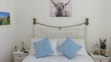 A neatly made bed with white bedding and two blue pillows, a brass bed frame, a small bedside table with a lamp, and a framed picture of a bull on the wall.