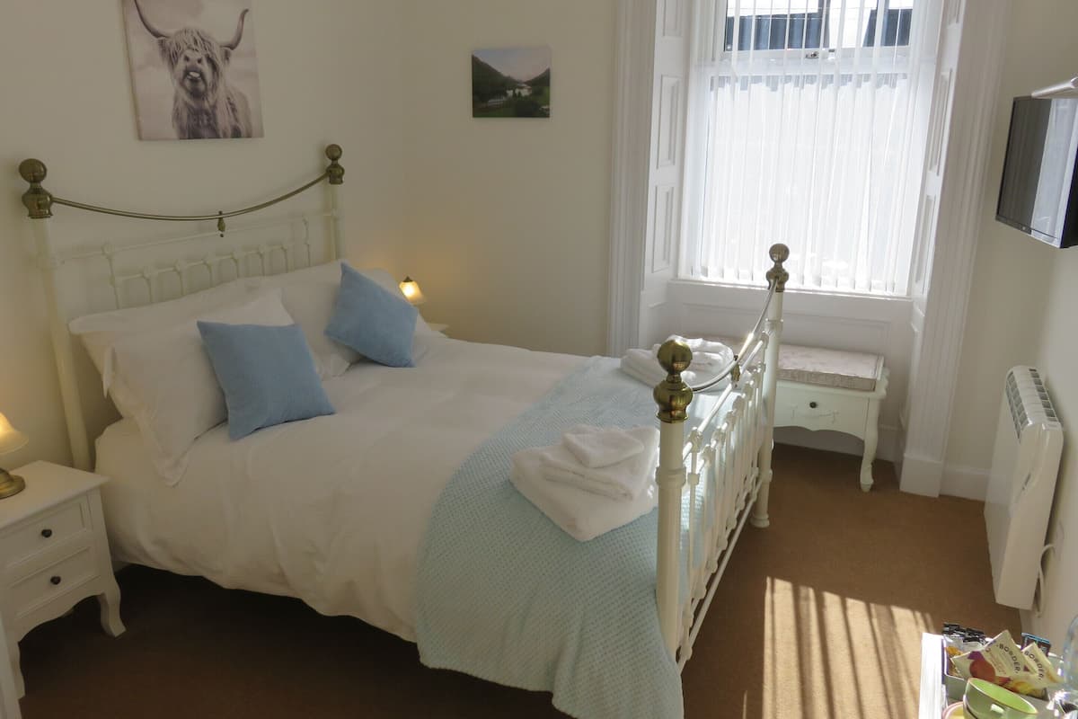 A neatly made bed with blue pillows, a bedside table with a lamp, and a window with sheer curtains.