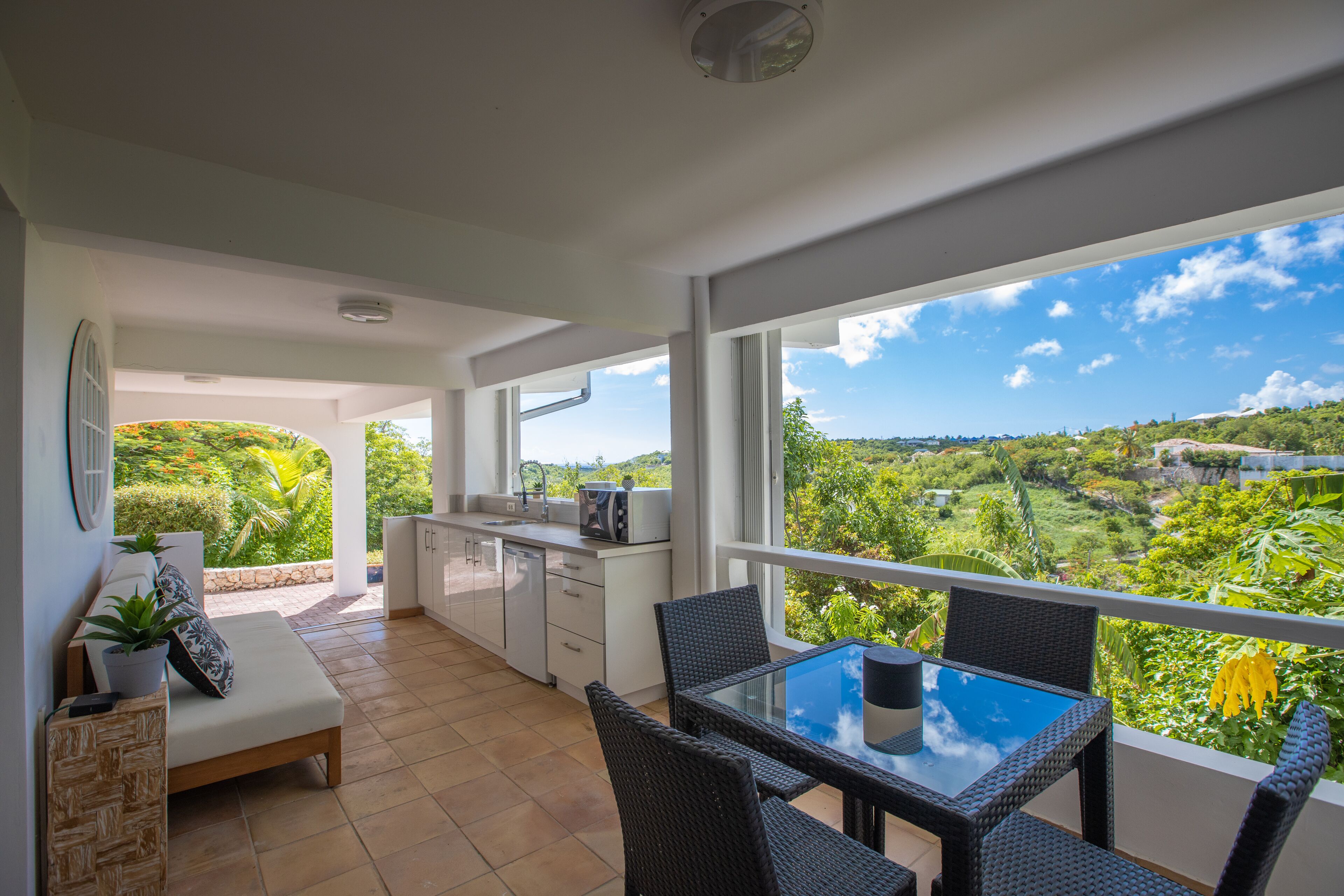 Une cuisine moderne avec des meubles blancs, un coin repas doté d’une table en verre et un balcon offrant une vue sur la verdure.