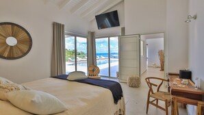 Une chambre avec un grand lit, un bureau et une télévision. Les fenêtres offrent une vue sur une piscine et une plage.