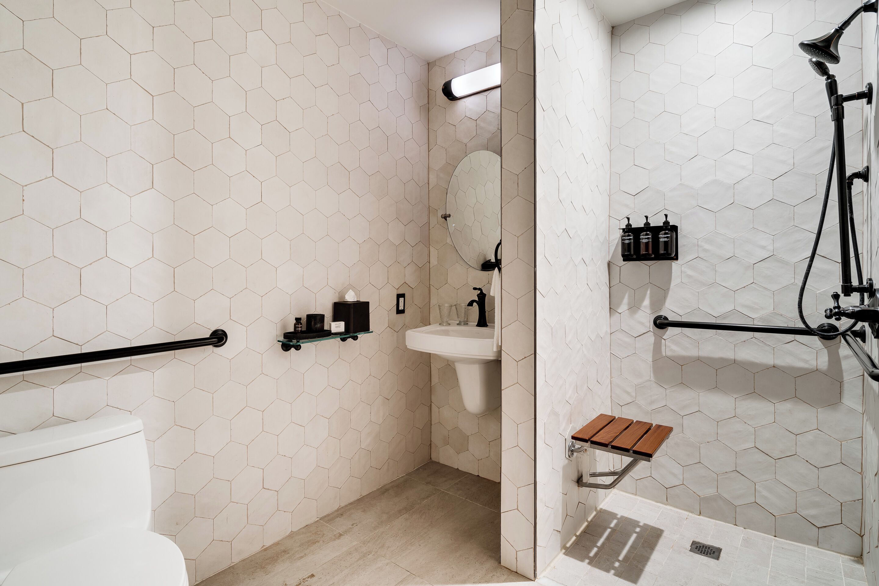 A bathroom with a hexagonal tile pattern, a white sink, a mirror, a handheld showerhead, and a wooden bench.