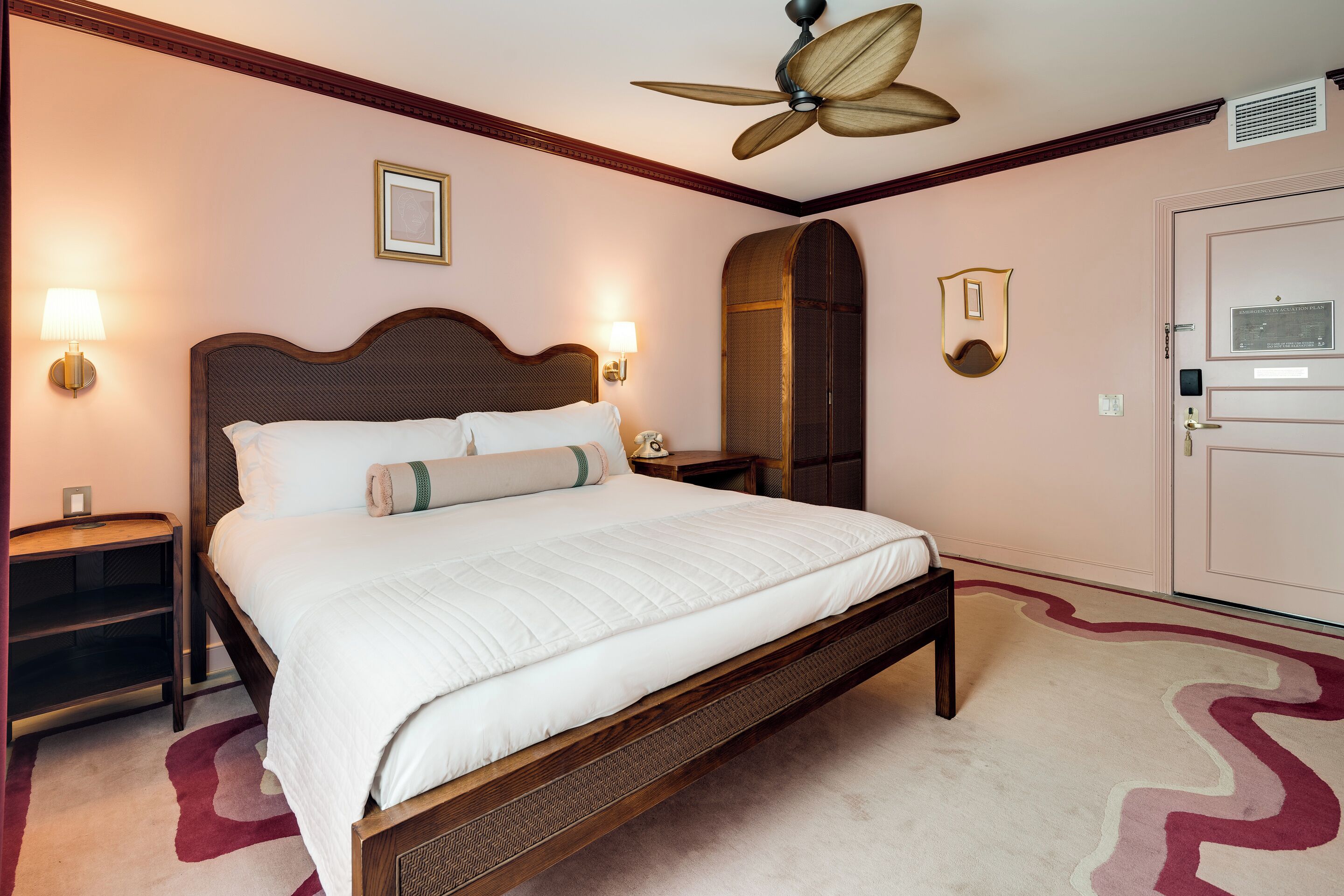Habitación de hotel con cama de madera, ventilador de techo, espejo adosado a la pared y puerta con placa.