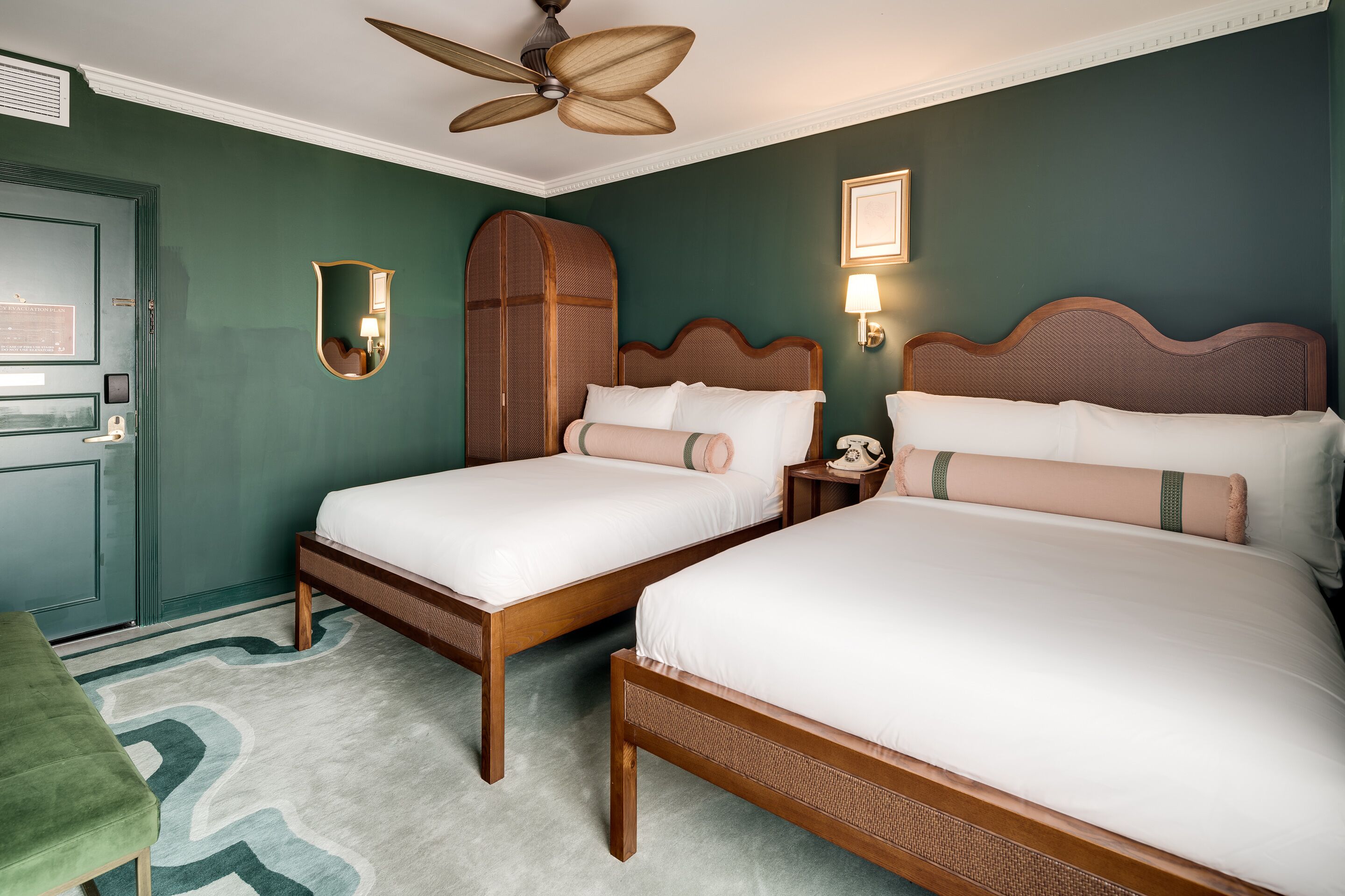 A hotel room with two wooden beds, a green accent wall, a ceiling fan, and a green accent chair.