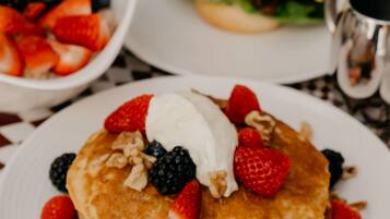 Desayuno con tortitas cubiertas de frutos rojos y una cucharada de crema, un sándwich y una taza de café sobre un mantel a cuadros.