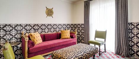 A room with a patterned wall, a red sofa, a green chair, a leopard print ottoman, and a window with curtains.