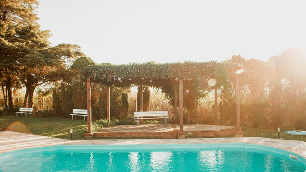 En swimmingpool med et pergola-område og bænke i en have.