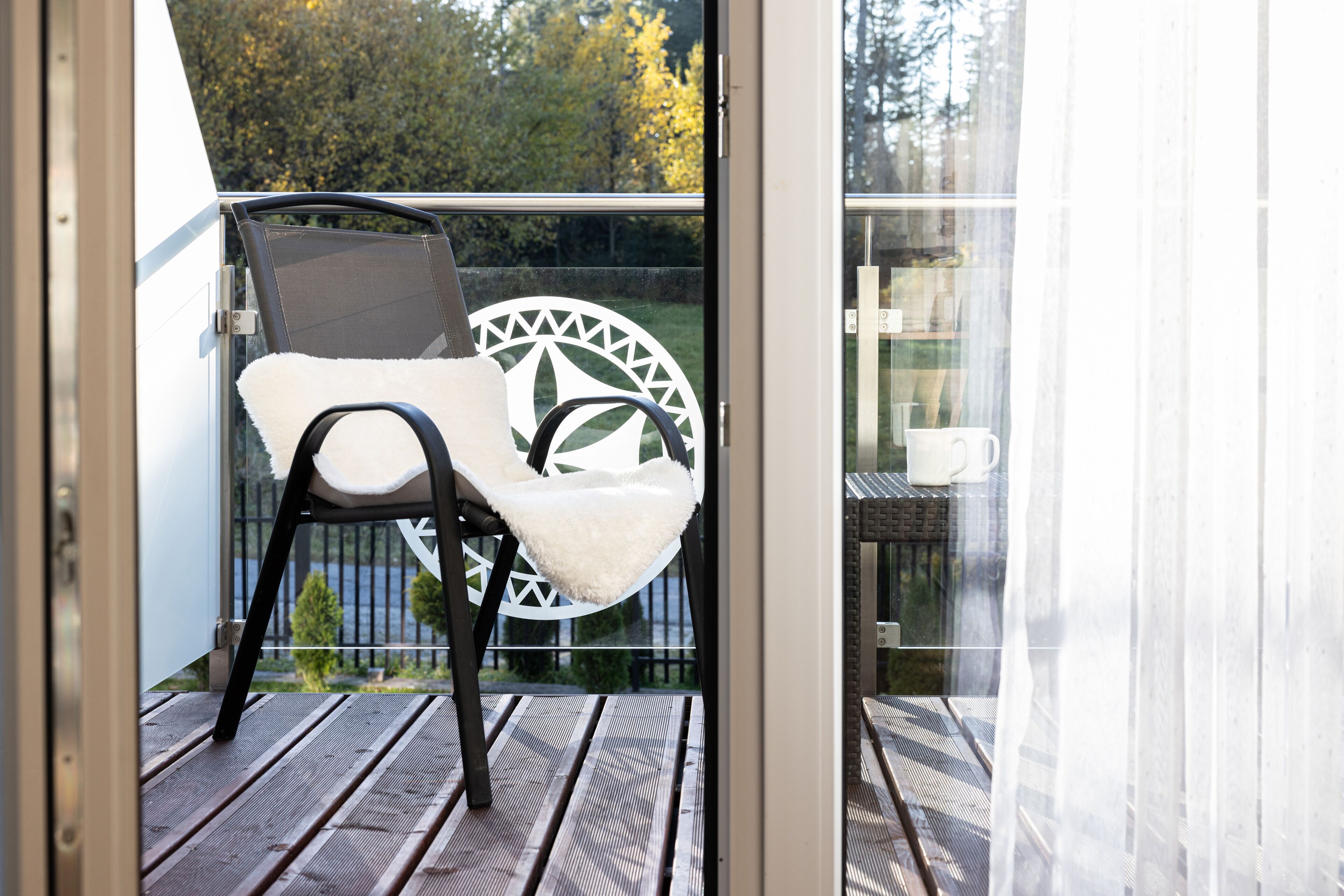 Un balcon avec une chaise et une table, une porte vitrée et une vue sur des arbres et une clôture.