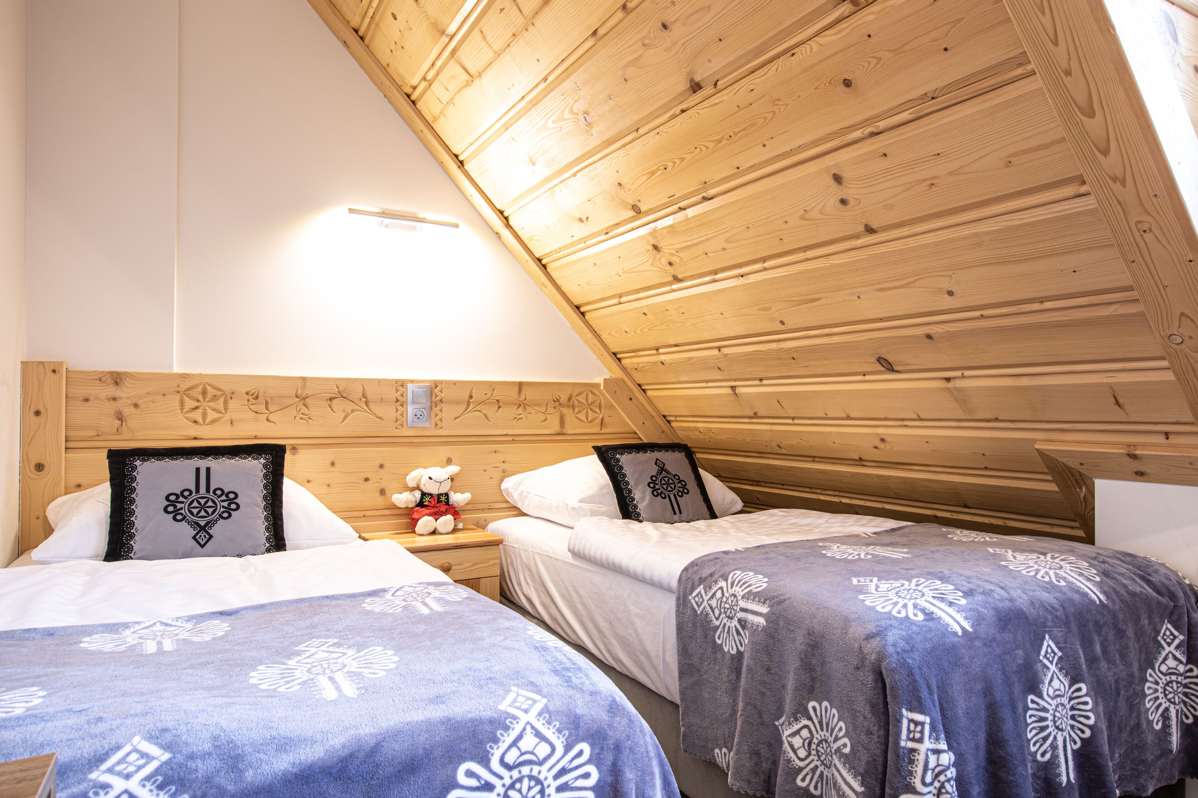 Une chambre chaleureuse avec deux lits, des poutres apparentes en bois au plafond et une table de chevet sur laquelle est posé un ours en peluche.
