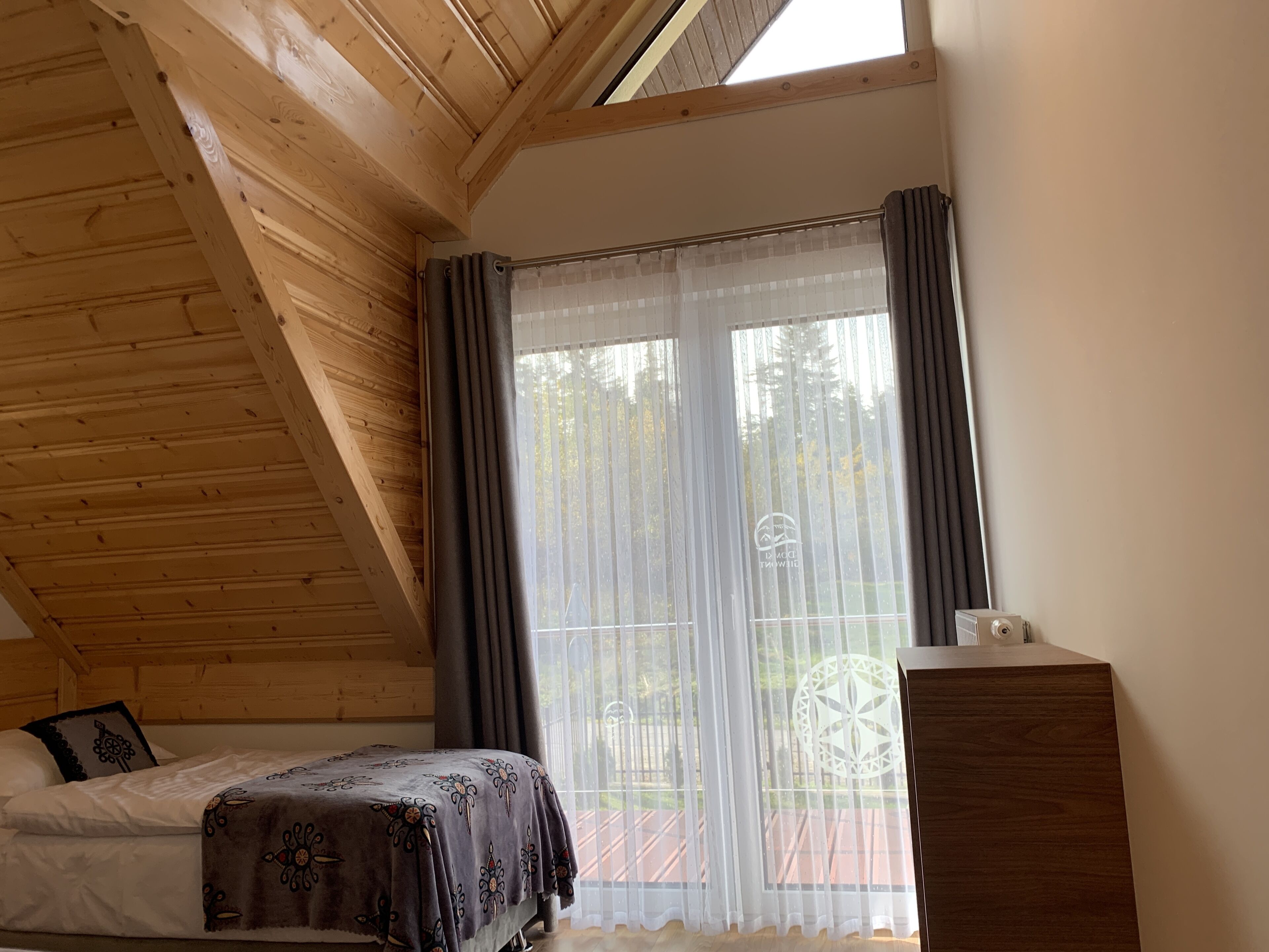 A bedroom with a wooden ceiling, a bed with a patterned blanket, a window with sheer curtains, and a small bedside table.