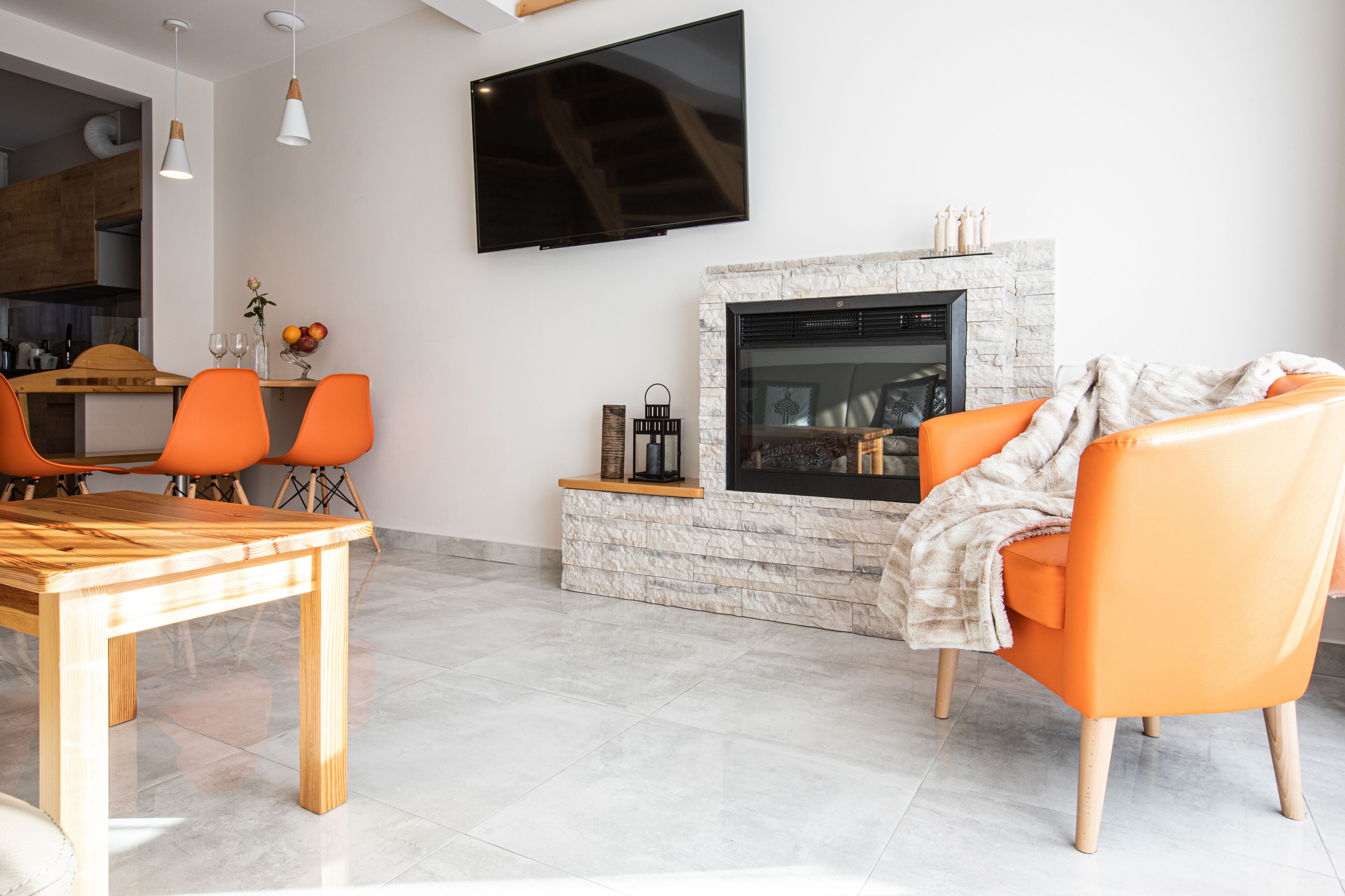 Un salon moderne avec une cheminée, un coin repas avec des chaises orange et un téléviseur fixé au mur.