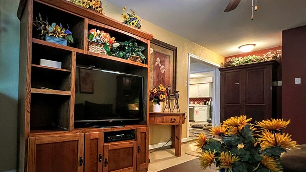 Een woonkamer met een houten entertainmentcentrum, een flatscreen-televisie en een bloemstuk op een bijzettafeltje.