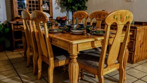 Een eetruimte met een houten tafel, gedekt voor zes personen, omringd door bijpassende stoelen. Achtergrond is een houten kast met glazen deuren, en een potplant staat ernaast.