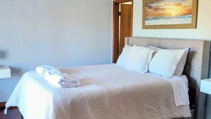 Una cama bien tendida con sábanas y almohadas blancas, un cuadro de un atardecer en la pared y una puerta de madera.