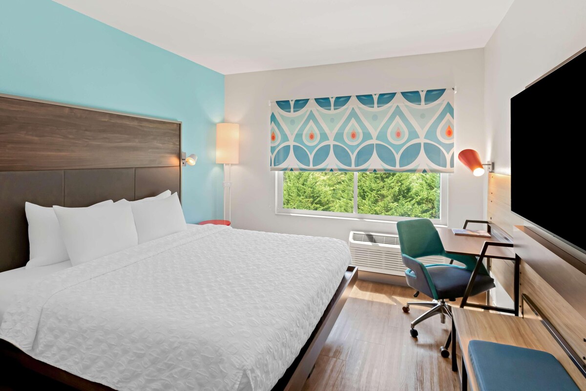A hotel room with a bed, a desk, a chair, a TV, a window with a view, and a patterned curtain.