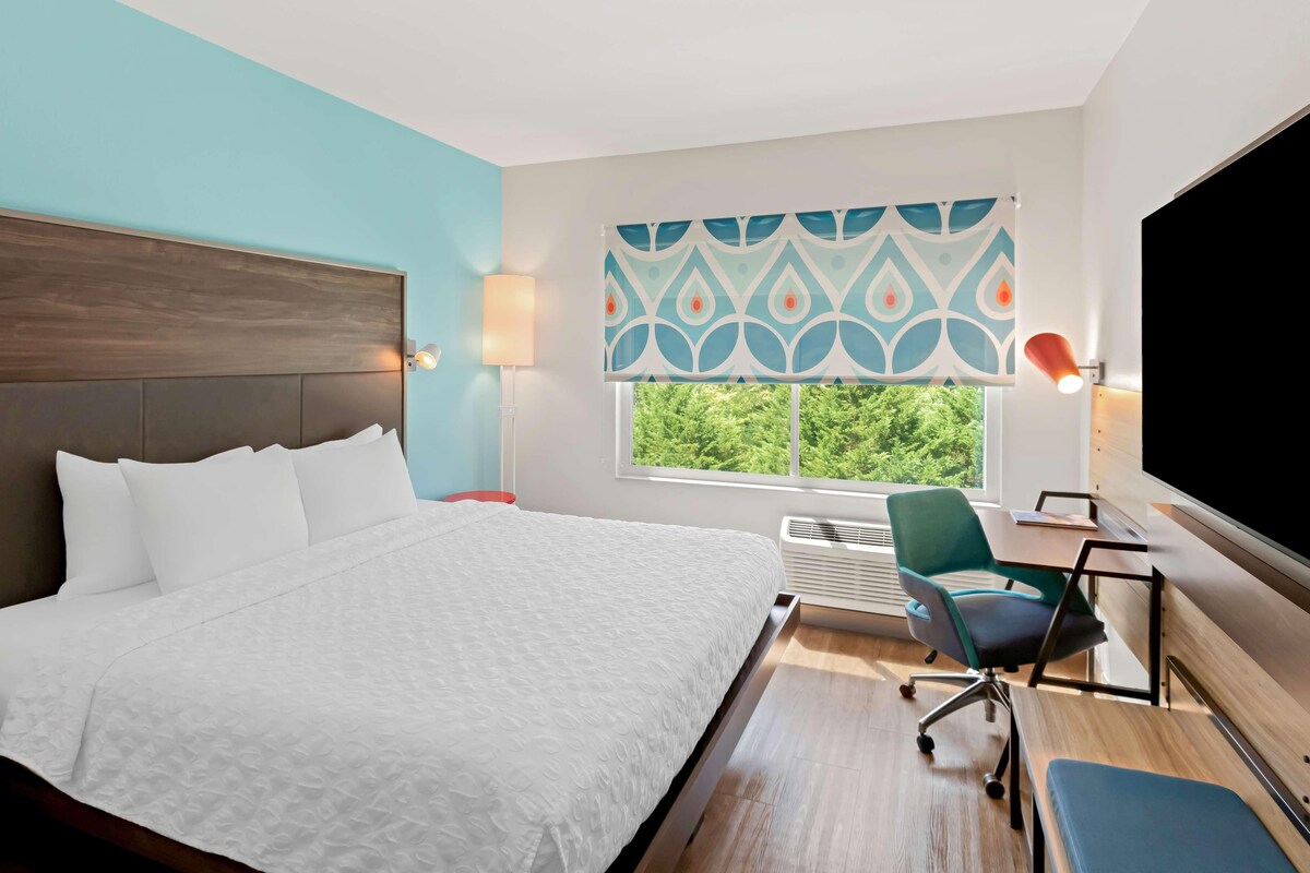 Quarto de hotel com uma cama, uma escrivaninha, uma cadeira, uma televisão, uma janela com vista e cortina estampada.