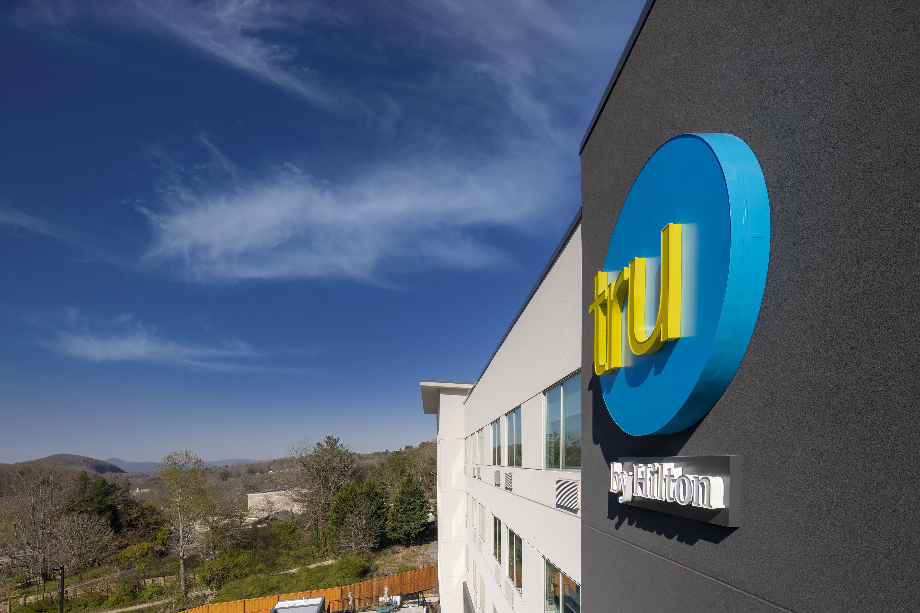 A modern building with a prominent blue and yellow logo, a sign indicating it's a Hilton hotel, and a view of a hilly landscape in the background.