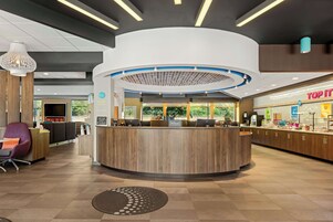 A modern hotel lobby with a central reception desk, seating area, and a 'Top It' counter.