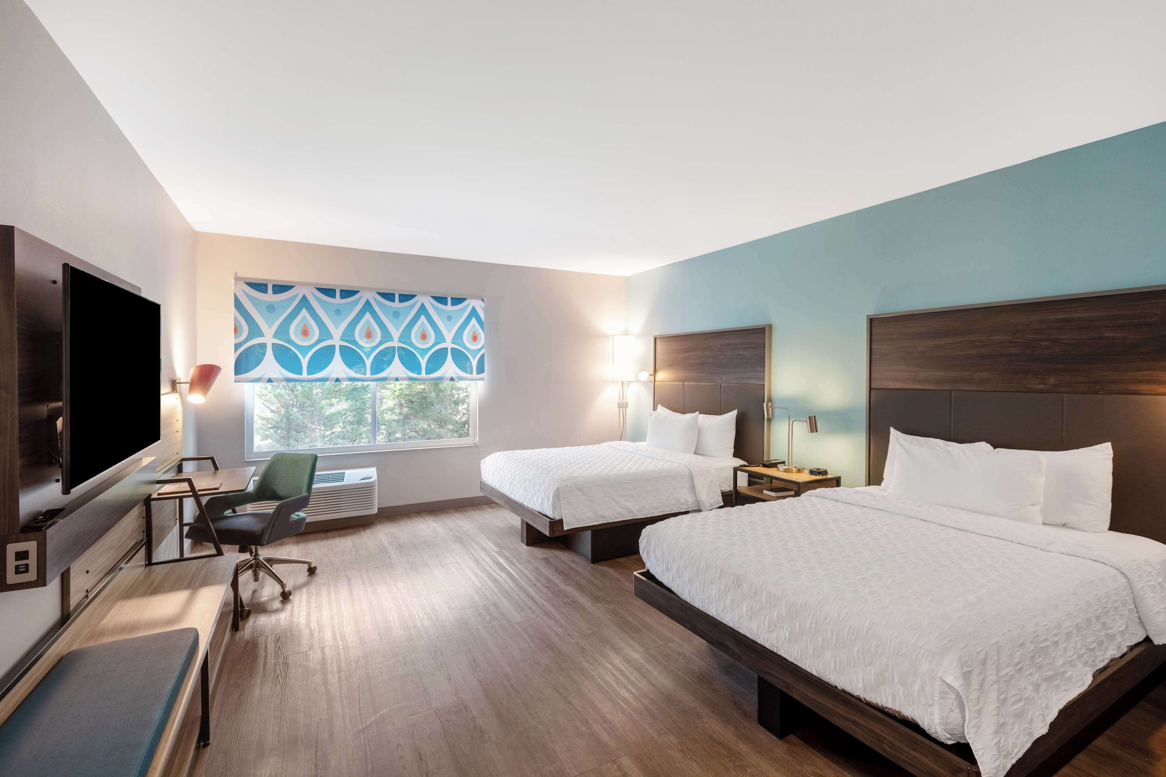 A hotel room with two beds, a desk, a chair, a TV, and a window with a blue patterned view.