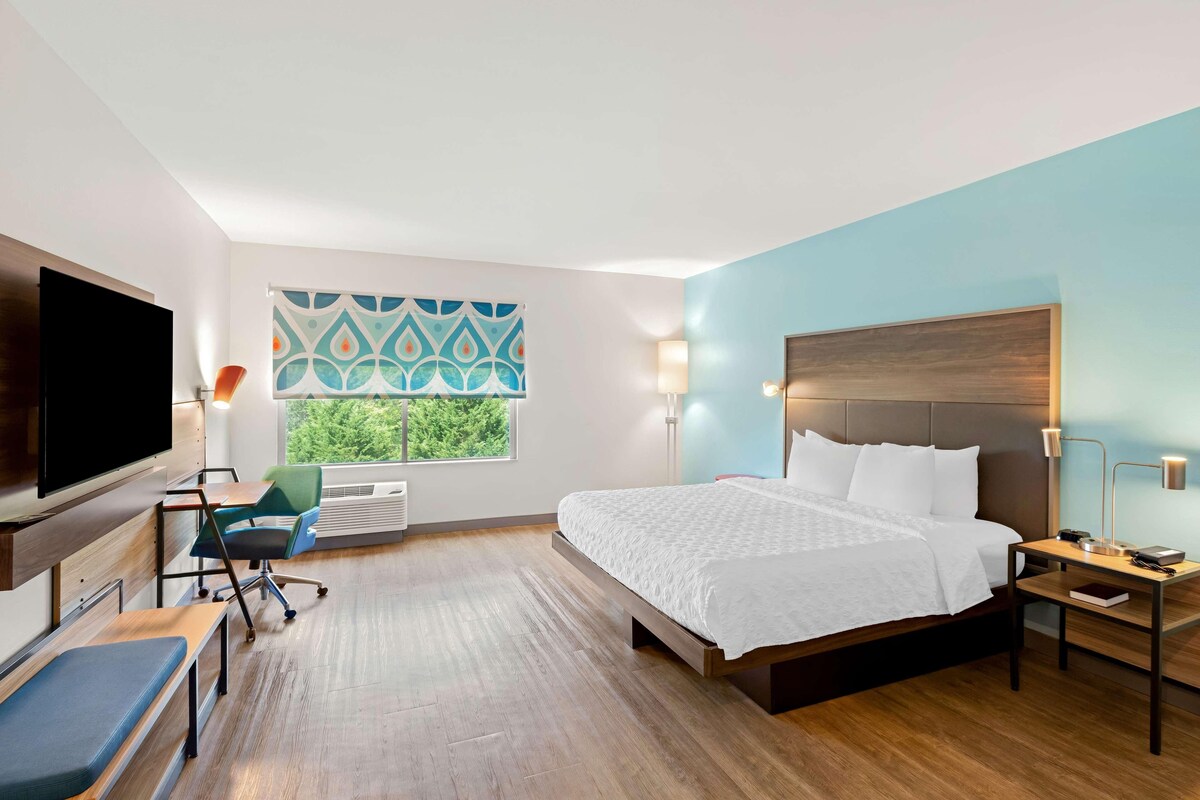 A hotel room with a bed, a desk, a chair, a TV, and a window with a view of greenery.