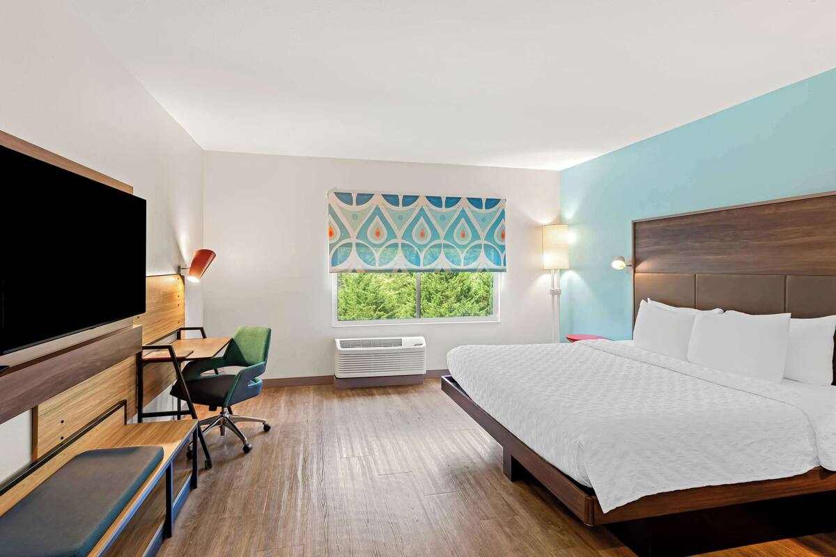 Ein Hotelzimmer mit Bett, Schreibtisch mit Stuhl, Fernseher, Fenster mit Blick ins Grüne und einer Wand-Klimaanlage.