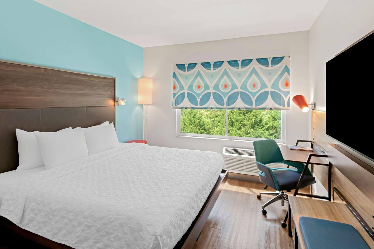 Ein Hotelzimmer mit Bett, Schreibtisch, Stuhl, Fernseher und einem Fenster mit Blick, dazu ein gemustertes Vorhang.