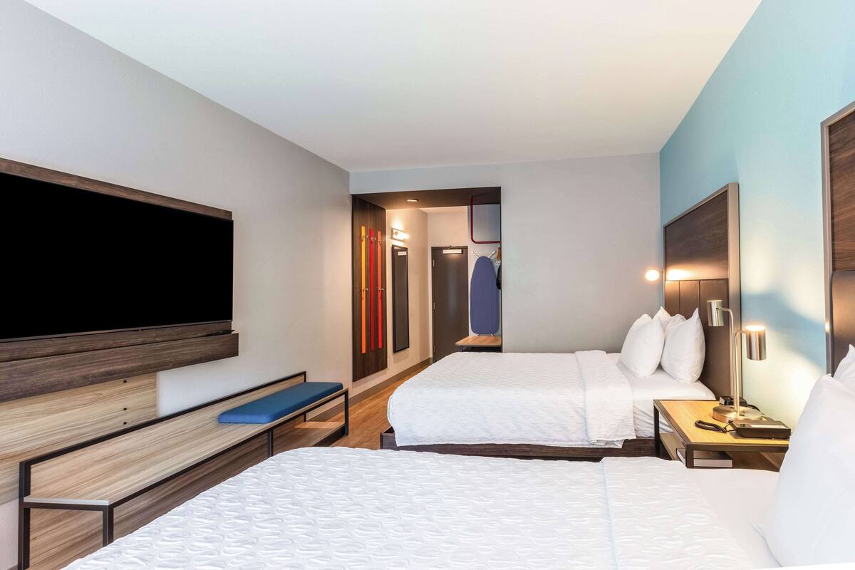 Ein Hotelzimmer mit zwei Betten, einem Fernseher, einer Sitzbank, einem Schreibtisch mit Lampe und einem Kleiderschrank.