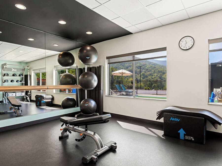 A gym with exercise equipment, a mirror, a clock, and a view of a mountainous landscape through large windows.