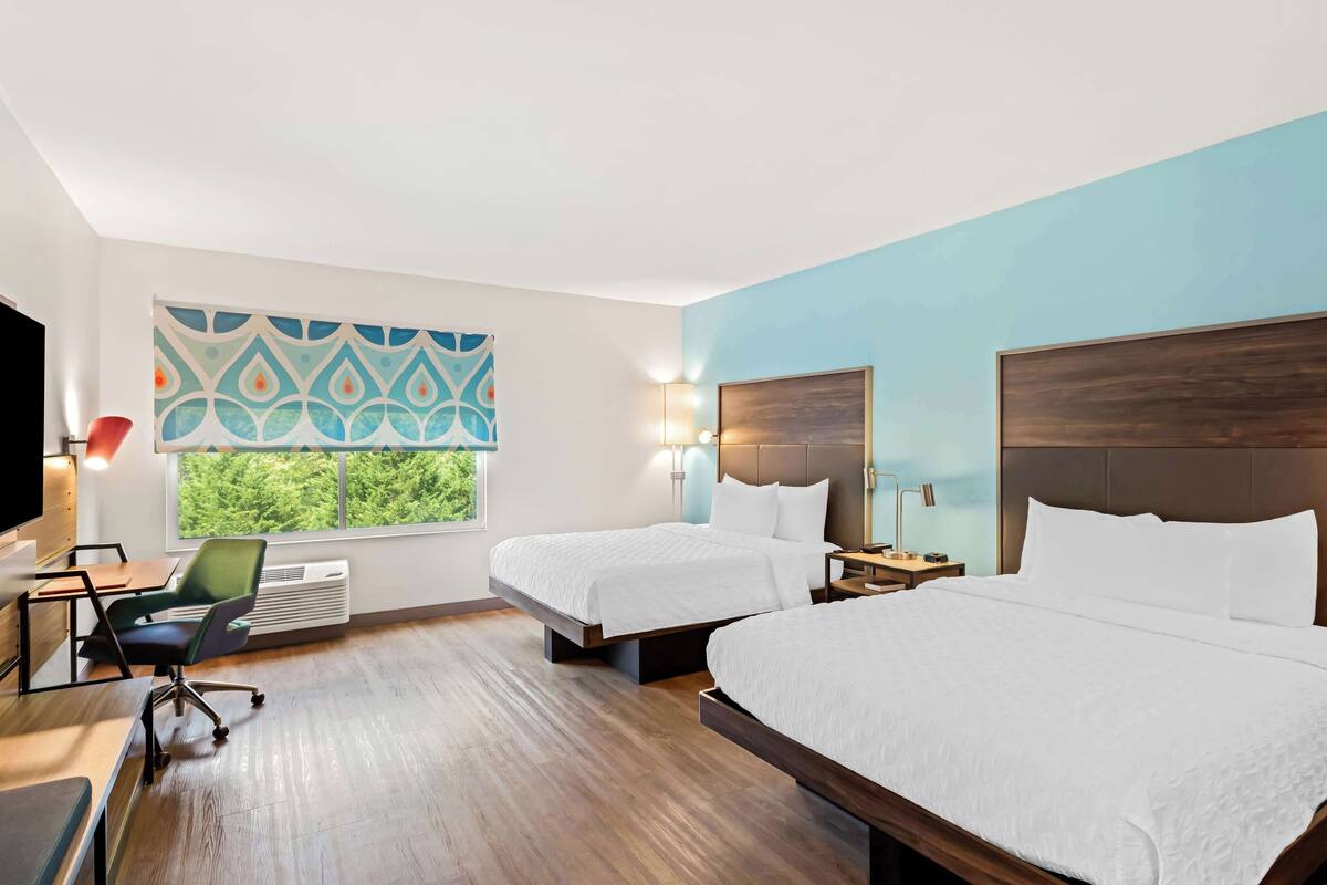 Ein Hotelzimmer mit zwei Betten, einem Schreibtisch, einem Stuhl, einem Fernseher und einem Fenster mit Blick auf Bäume.