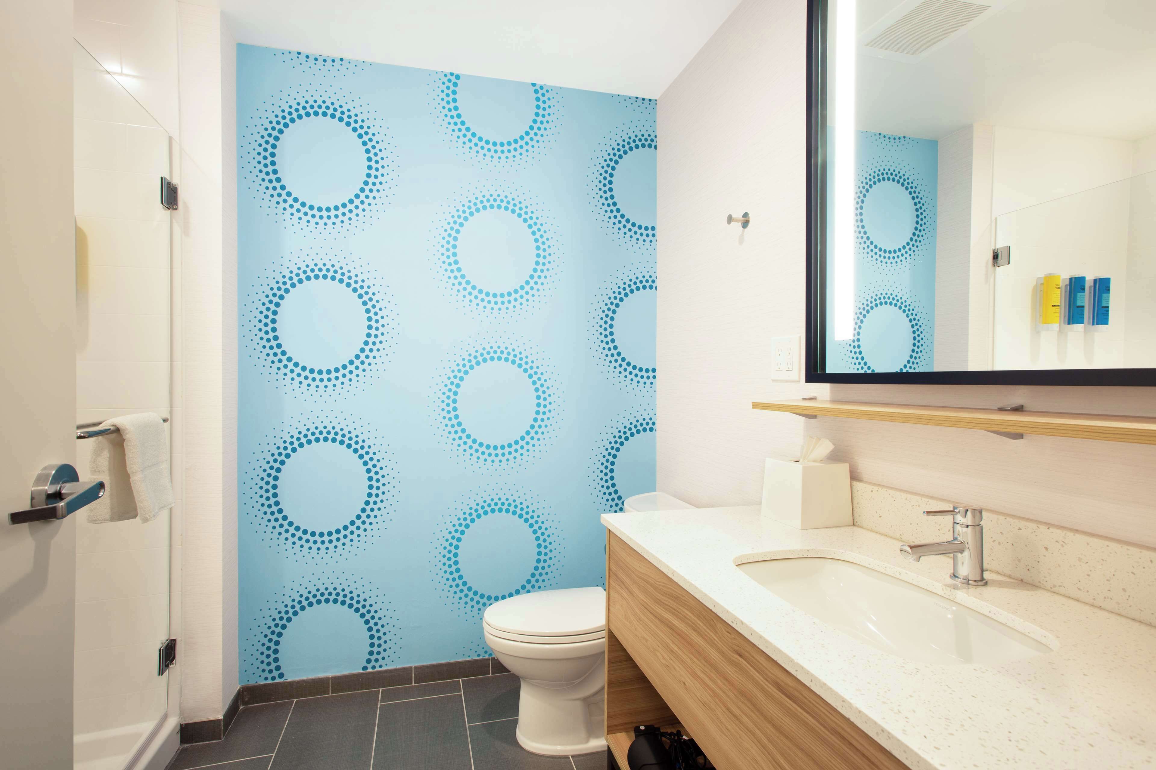 A modern bathroom with a blue circular pattern on the wall, a white toilet, a wooden vanity with a sink, and a mirror above the vanity.