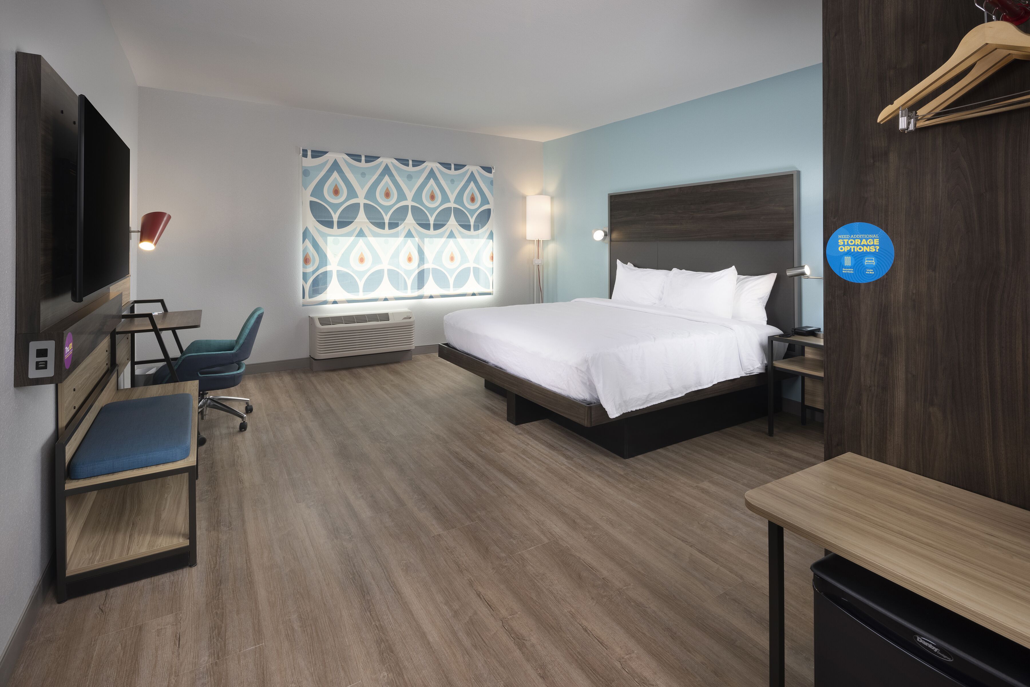 A hotel room with a bed, desk, chair, TV, and a window with patterned curtains.