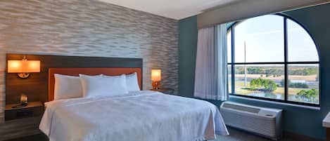 A hotel room with a large bed, two bedside lamps, a view of a landscape through a large window, and a wall-mounted air conditioner.