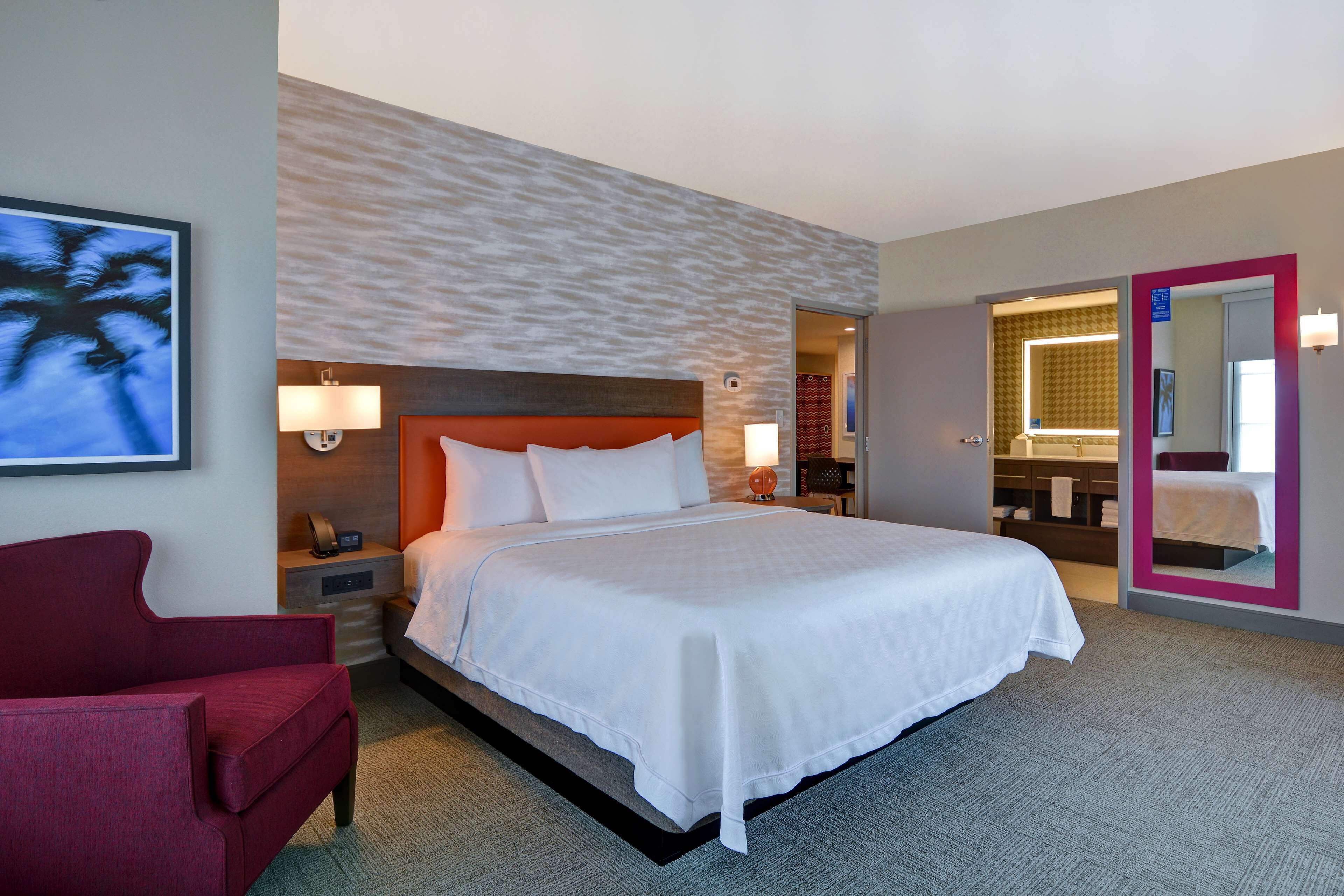 Une chambre d’hôtel avec un grand lit, un fauteuil rouge, un téléviseur fixé au mur et une salle de bain visible à travers une porte ouverte.